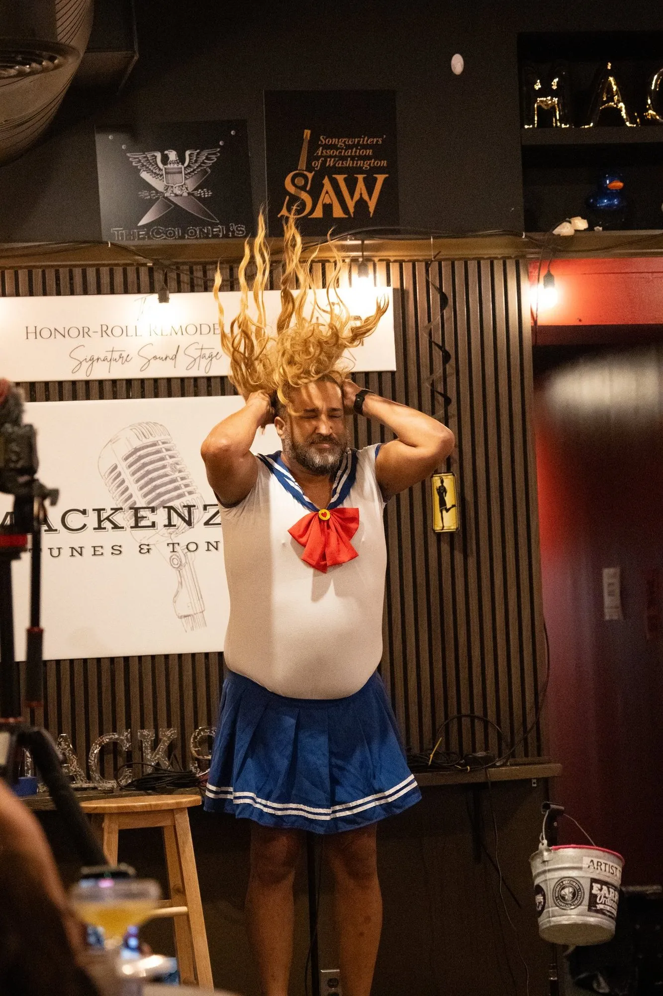 A performer in a sailor costume flipping a blonde wig on stage